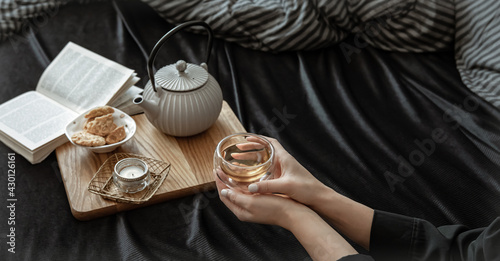 Cozy composition with a cup of tea in female hands, cookies and a book in bed.