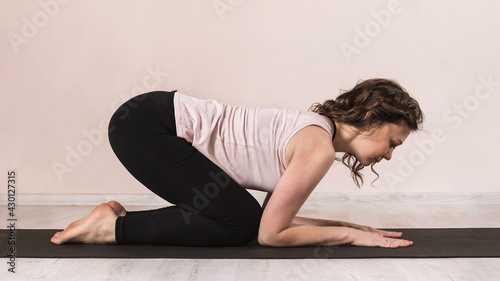 A woman in sportswear practicing yoga performs the bitilasana exercise, cat pose, on a mat near the wall