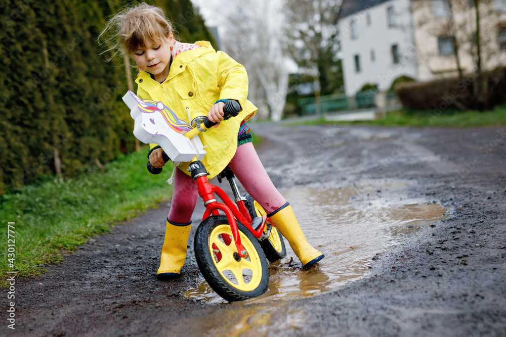 Little toddler girl wearing yellow rain gum boots, running with balance bike during sleet. Happy child driving, biking with bicycle into puddle, splashing water, outdoor activity. Happiness, childhood Little toddler girl wearing yellow rain gum boots, running with balance bike during sleet. Happy child driving, biking with bicycle into puddle, splashing water, outdoor activity. Happiness, childhood