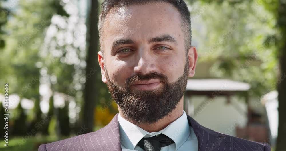 Happy successful mature bearded businessman wearing formal suit winking to camera and smiling, posing at public park Happy successful mature bearded businessman wearing formal suit winking to camera and smiling, posing at public park