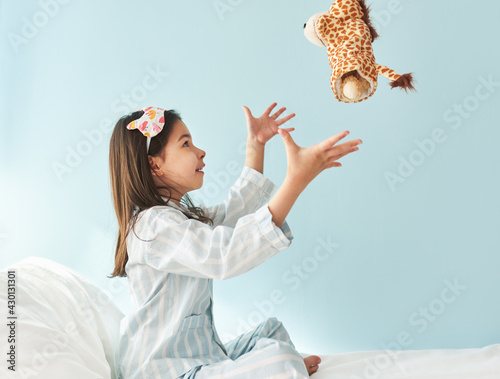 Sied view of cute little girl in blue pajama in white stripes, sleeping mask on head, smiling, and playing with toy giraffe in the bed. Kid has joyful expression in the morning.