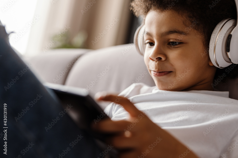Black curly boy in headphones using tablet computer while sitting on sofa Black curly boy in headphones using tablet computer while sitting on sofa