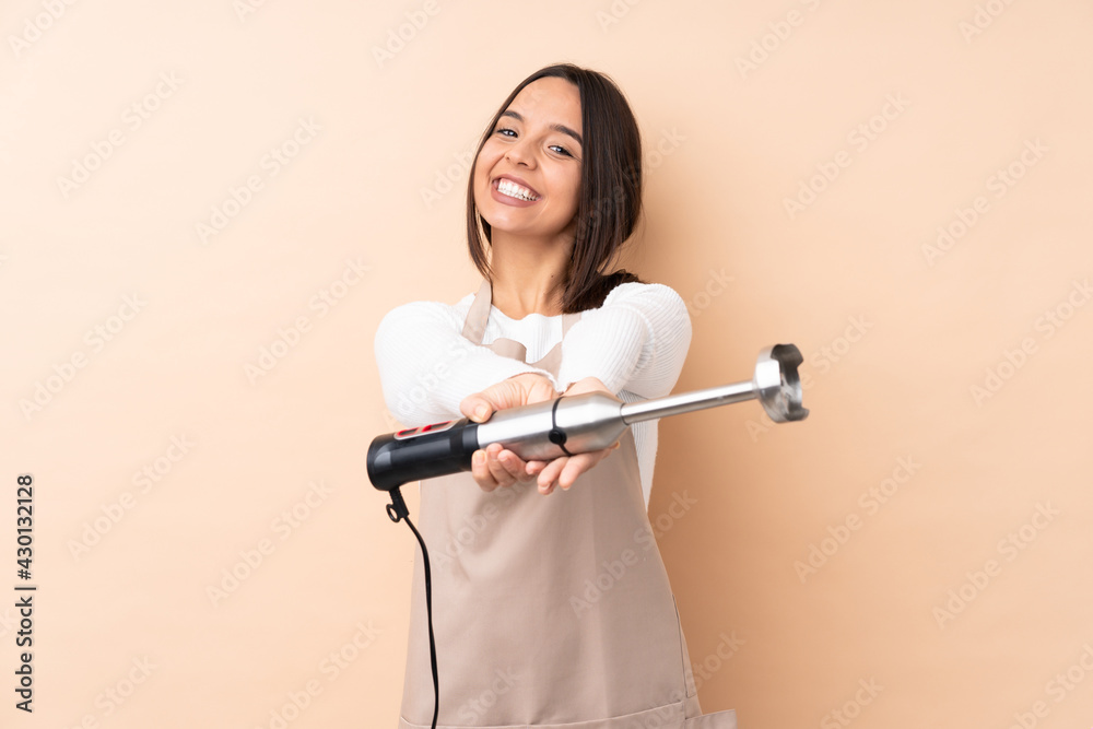 Young brunette girl using hand blender over isolated background holding copyspace imaginary on the palm to insert an ad Young brunette girl using hand blender over isolated background holding copyspace imaginary on the palm to insert an ad