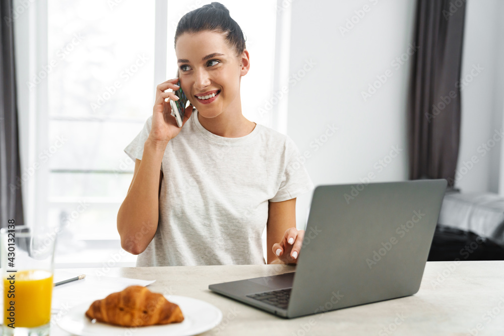Smiling woman talking on cellphone and using laptop while having breakfast Smiling woman talking on cellphone and using laptop while having breakfast