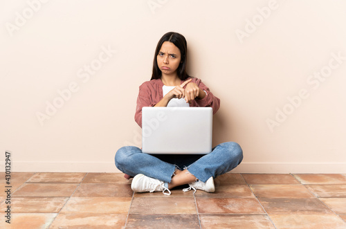 Young mixed race woman with a laptop sitting on the floor making the gesture of being late © luismolinero