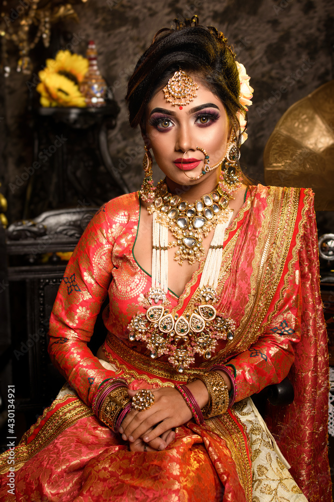 Stunning Indian bride in luxurious bridal costume with makeup and heavy  jewellery is sitting in a chair in with classic vintage interior in studio  lighting. Wedding lifestyle and fashion. Photos