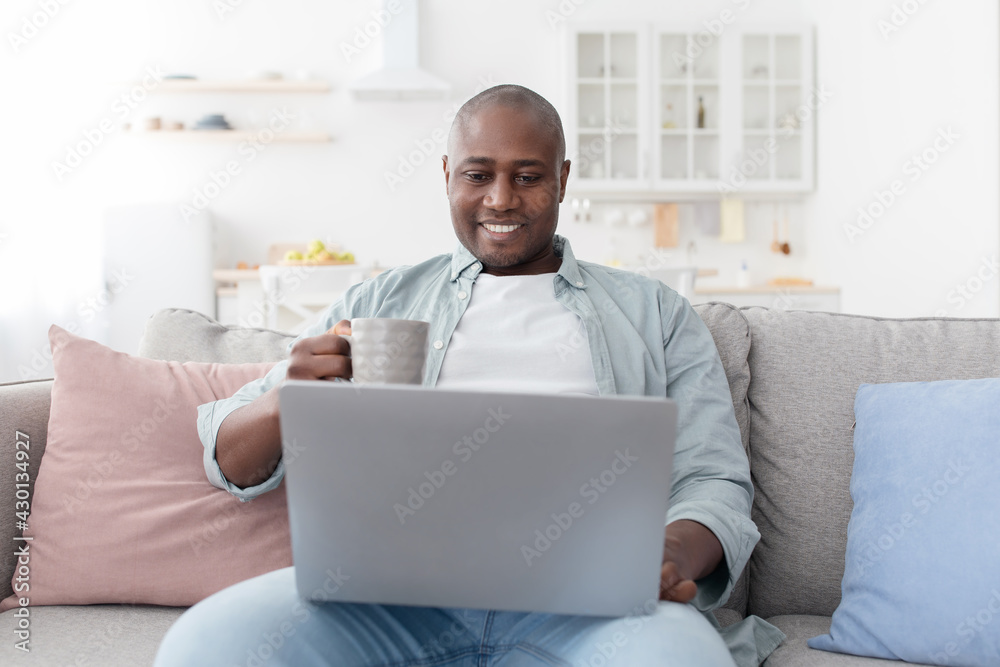 Reading morning news. Happy african american man using laptop and drinking hot coffee, resting on sofa Reading morning news. Happy african american man using laptop and drinking hot coffee, resting on sofa