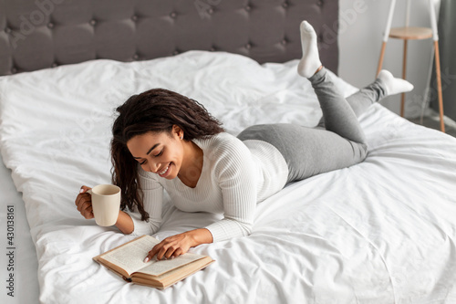 Black woman reading in bed, holding cup of hot coffee