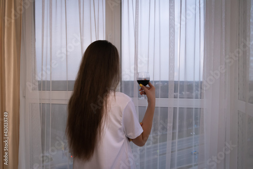 Young woman with long dark hair and a white oversize T-shirt is standing at the window with a glass of red wine, looking out into the distance of the city. Photo from the back photo