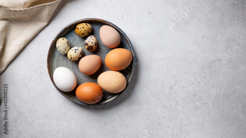 Different types of eggs in bowl on gray background. Chicken, guinea fowl and quail eggs on plate. Top view, copy space.