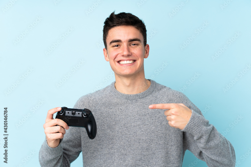 Teenager caucasian man playing with a video game controller isolated on blue background and pointing it Teenager caucasian man playing with a video game controller isolated on blue background and pointing it
