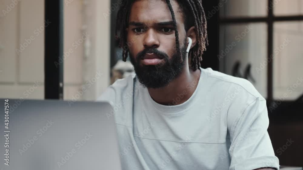 A serious african american man with earbuds is using his laptop while sitting on the street A serious african american man with earbuds is using his laptop while sitting on the street