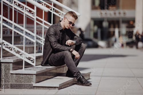 Young fashion man looks at his watch sitting in a city street