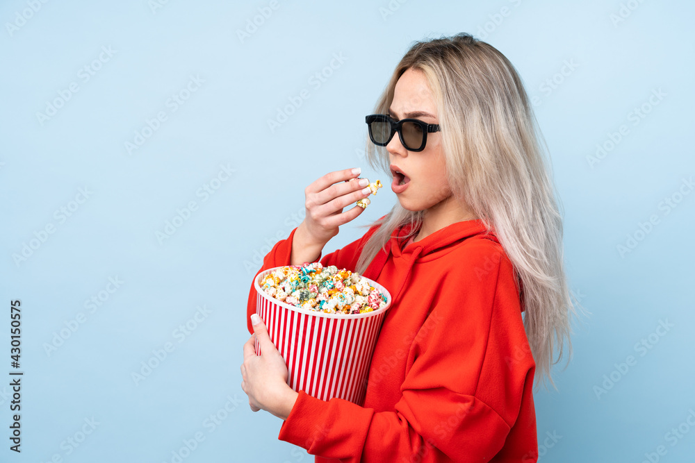 Teenager girl over isolated blue background with 3d glasses and holding a big bucket of popcorns while looking side