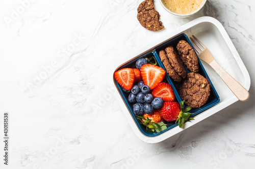 Lunch box with fruits and cookies on white marble background, top view. Takeaway healthy snack container.