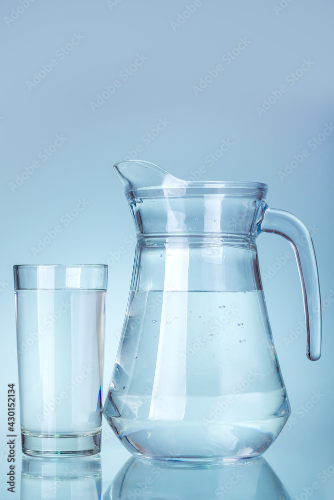 A clear glass jug with a handle and a spout filled with drinking water stands by a glass of liquid on a cool blue background kitchen utensil. A clear glass jug with a handle and a spout filled with drinking water stands by a glass of liquid on a cool blue background kitchen utensil.