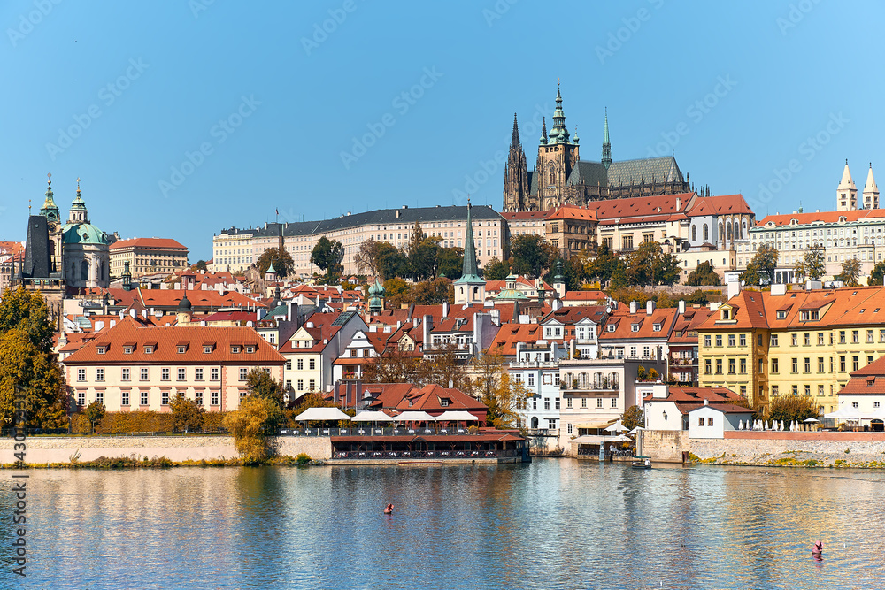 St. Vitus Cathedral and Prague Castle with orange roofs of historic buildings of Mala Strana reflected in river Vltava on a bright Summer day in Prague, Czech Republic.