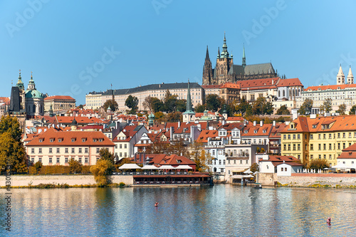 St. Vitus Cathedral and Prague Castle with orange roofs of historic buildings of Mala Strana reflected in river Vltava on a bright Summer day in Prague, Czech Republic.