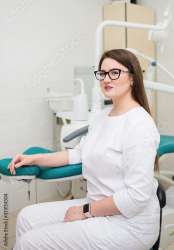 a woman doctor with brown hair glasses and a white suit is sitting in the office near the couch