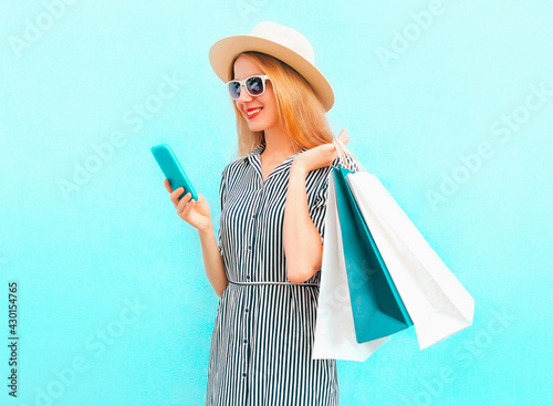 Portrait of happy smiling young woman with smartphone and shopping bags wearing a summer straw hat, striped dress on a blue background