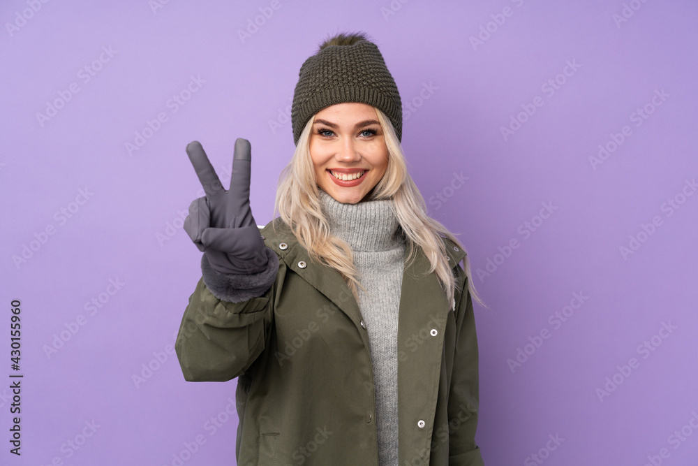 Teenager blonde girl with winter hat over isolated purple background smiling and showing victory sign Teenager blonde girl with winter hat over isolated purple background smiling and showing victory sign