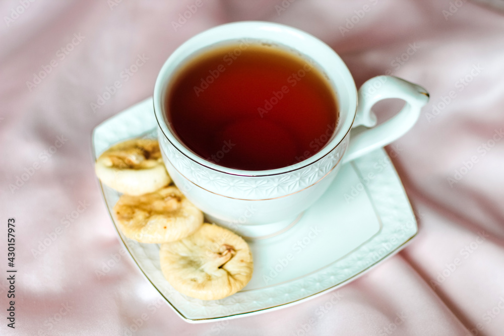 White cup with tea with a saucer, next to dried dates dessert on a silk tablecloth. White cup with tea with a saucer, next to dried dates dessert on a silk tablecloth.