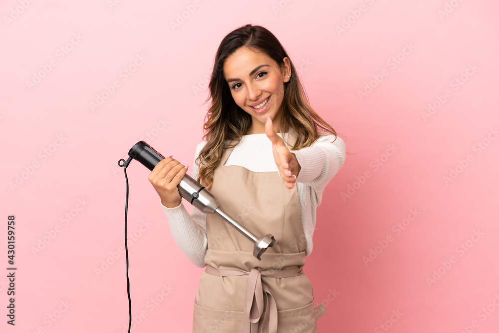 Young woman using hand blender over isolated pink background shaking hands for closing a good deal Young woman using hand blender over isolated pink background shaking hands for closing a good deal