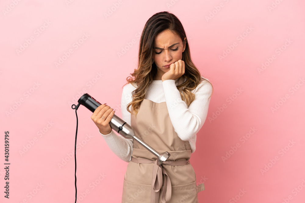 Young woman using hand blender over isolated pink background having doubts