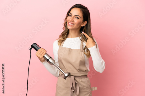 Young woman using hand blender over isolated pink background giving a thumbs up gesture