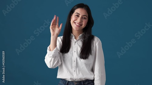 A smiling korean woman is waving hand showing goodbye gesture standing isolated over blue wall in studio photo