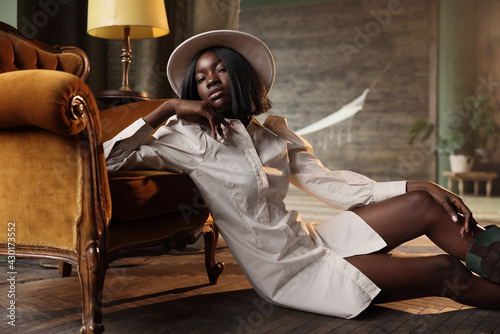 Photoshooting of a beautiful dark-skinned girl in the studio sitting on the floor near a chair, dressed in a beige dress, hat and boots. She poses in the camera.