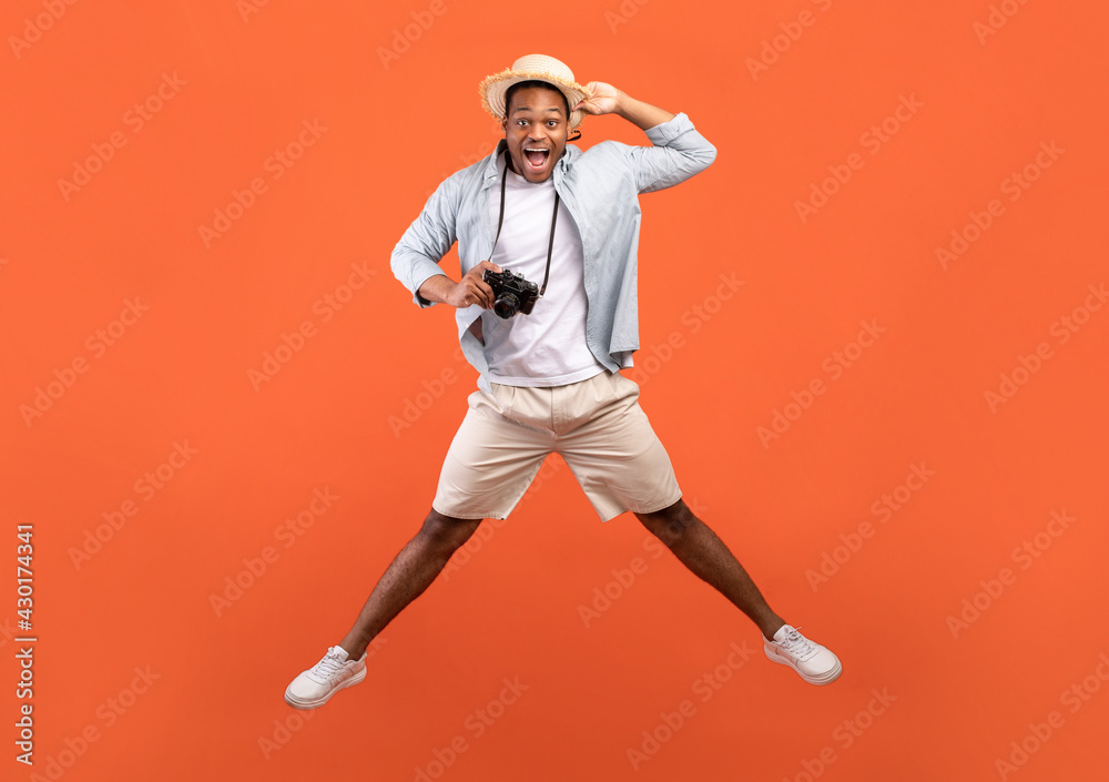 Full length of excited black man in summer outfit holding professional camera and jumping on orange studio background