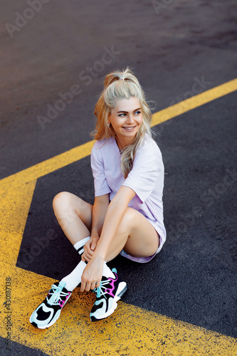 Image of beautiful stylish woman sitting on street with legs crossed smiling on summer day. Cheerful girl in sport wear is sitting in the middle of the road. Sporty, blonde woman with curly hair.