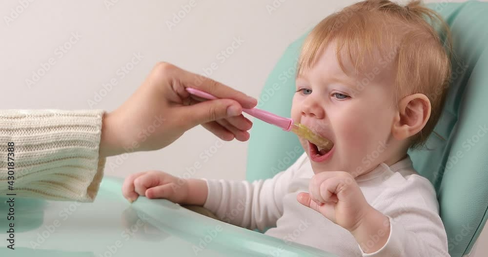Closeup of young baby in baby chair and mother hand with a spoon, while she is feeding the baby, making funny grimaces. Closeup of young baby in baby chair and mother hand with a spoon, while she is feeding the baby, making funny grimaces.