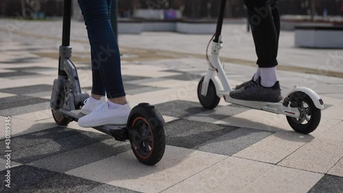 A man and a woman ride electric scooters. In the frame, the legs and footboards of scooters.