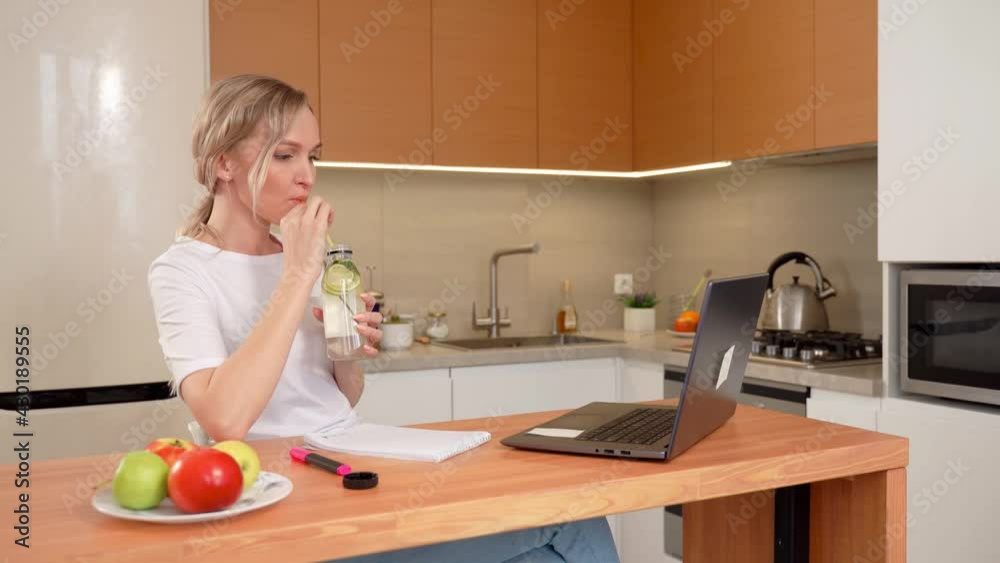 The blonde is sitting at the table in the kitchen, drinking lemonade from a straw, communicating via video link looking at a laptop. Pretty business girl using laptop in the kitchen The blonde is sitting at the table in the kitchen, drinking lemonade from a straw, communicating via video link looking at a laptop. Pretty business girl using laptop in the kitchen