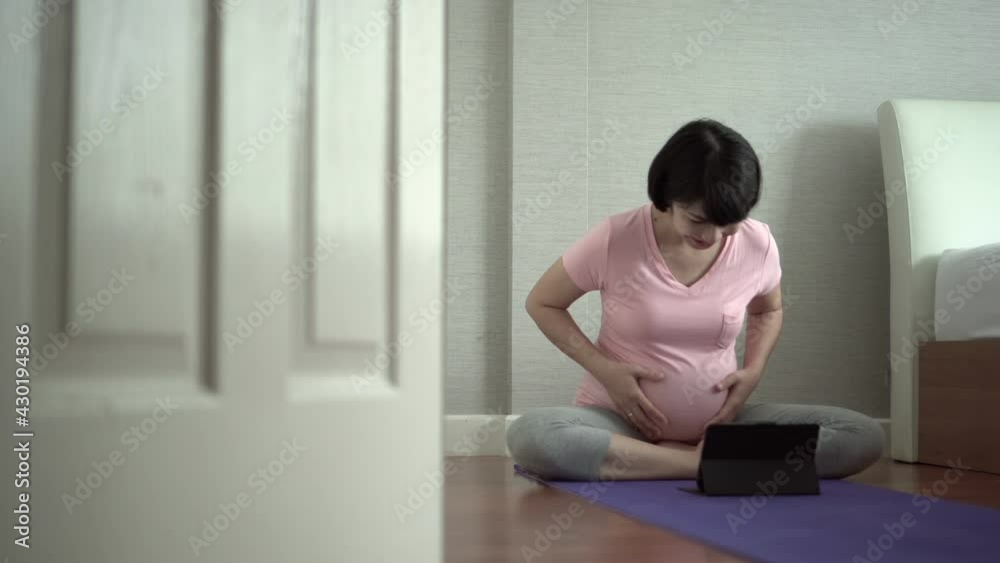 Happy Smiling Asian pregnant woman sitting on floor and touching her belly while doing video call with family on digital tablet Happy Smiling Asian pregnant woman sitting on floor and touching her belly while doing video call with family on digital tablet