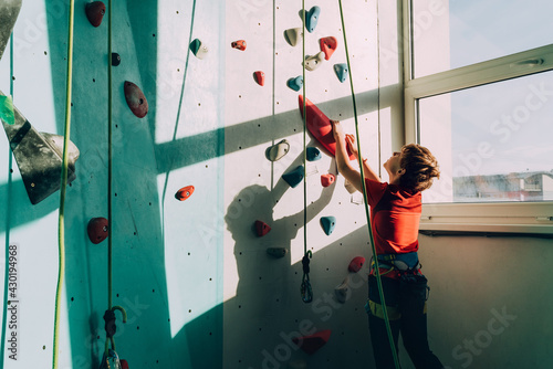 Teenager boy at indoor climbing wall hall starting climbing. Boy using climbing harness and chalk powder when somebody belaying him from floor. Active teenager time spending concept image.
