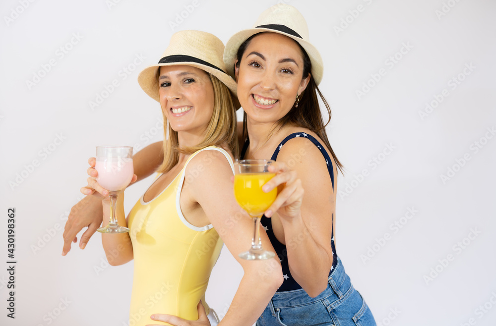 Two cheerful young women girls friends in summer clothes with straw hat, holding beverages isolated over white background.