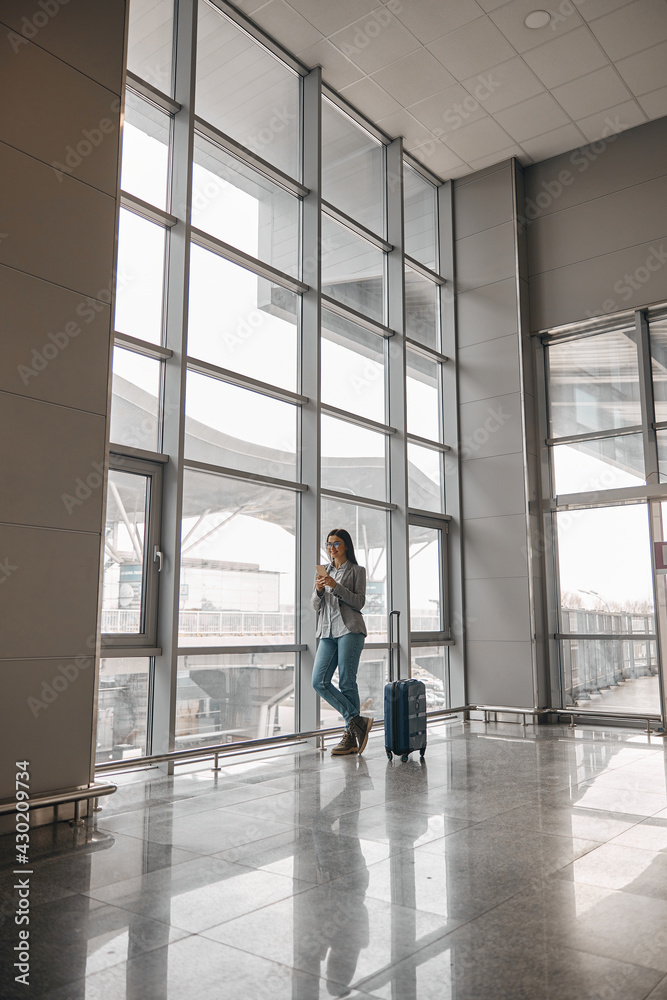 Caucasian woman is standing alone near airport terminal panoramic windows with luggage