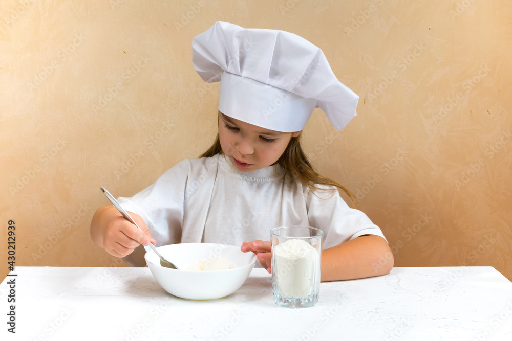 A little girl dressed as a cook kneads the dough. Cooking child lifestyle concept. The kid loves, has fun, studies and plays in the kitchen A little girl dressed as a cook kneads the dough. Cooking child lifestyle concept. The kid loves, has fun, studies and plays in the kitchen