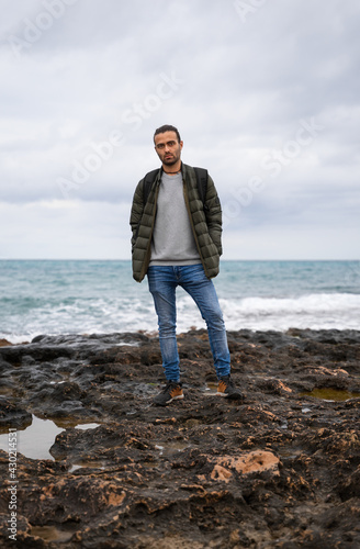 Man posing in the sea with attitude and a backpack. In a sea of rocks surrounded by puddles on a cloudy day. He is wearing a backpack, jeans, sweater and green jacket. His hands are in his pockets.