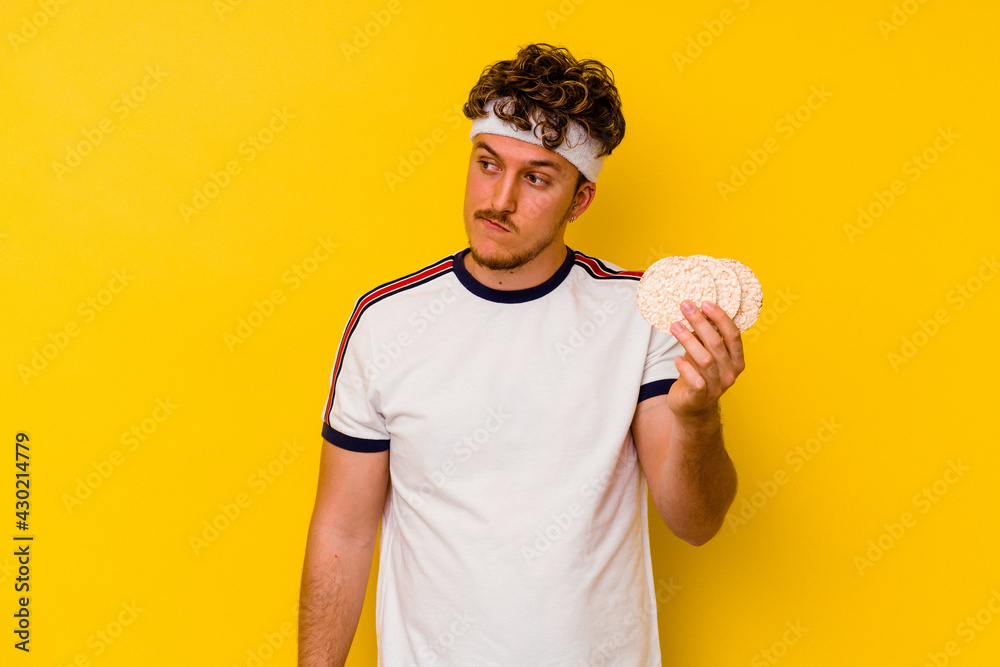 Young sport caucasian man eating a rice cake isolated on yellow background confused, feels doubtful and unsure. Young sport caucasian man eating a rice cake isolated on yellow background confused, feels doubtful and unsure.