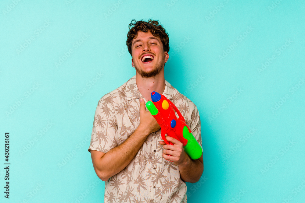 Young caucasian man holding a water gun isolated on blue background laughs out loudly keeping hand on chest. Young caucasian man holding a water gun isolated on blue background laughs out loudly keeping hand on chest.
