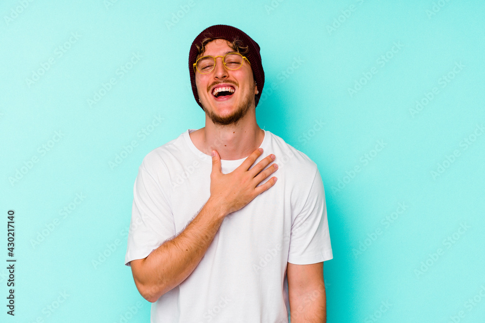 Young caucasian man wearing a wool hat isolated on blue background laughs out loudly keeping hand on chest. Young caucasian man wearing a wool hat isolated on blue background laughs out loudly keeping hand on chest.