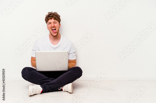 Young caucasian man sitting on the floor holding on laptop isolated on white background funny and friendly sticking out tongue.