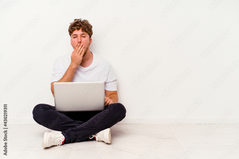 Young caucasian man sitting on the floor holding on laptop isolated on white background yawning showing a tired gesture covering mouth with hand. Young caucasian man sitting on the floor holding on laptop isolated on white background yawning showing a tired gesture covering mouth with hand.
