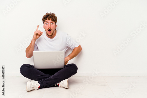 Young caucasian man sitting on the floor holding on laptop isolated on white background having an idea, inspiration concept.