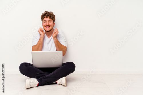 Young caucasian man sitting on the floor holding on laptop isolated on white background raising fist, feeling happy and successful. Victory concept.
