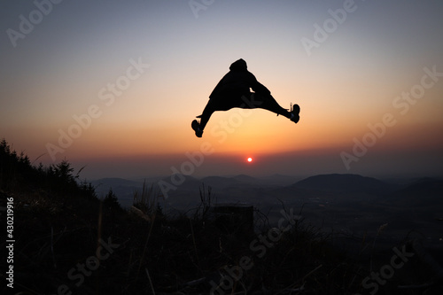 Silhouette of a teenager and athlete jumping on a tree stump and doing a straddle vault, splits at sunset in the European mountains. Training in natural conditions. Self weight training. Flexibility